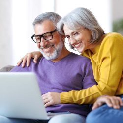 Beautiful mature family couple looking at laptop with smile on face while spending great time together at home on retirement, happy retired husband and wife watching funny video on computer